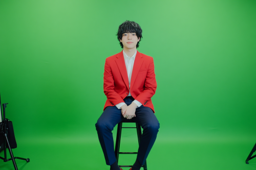 Japanese man in red blazer sitting on stool.
