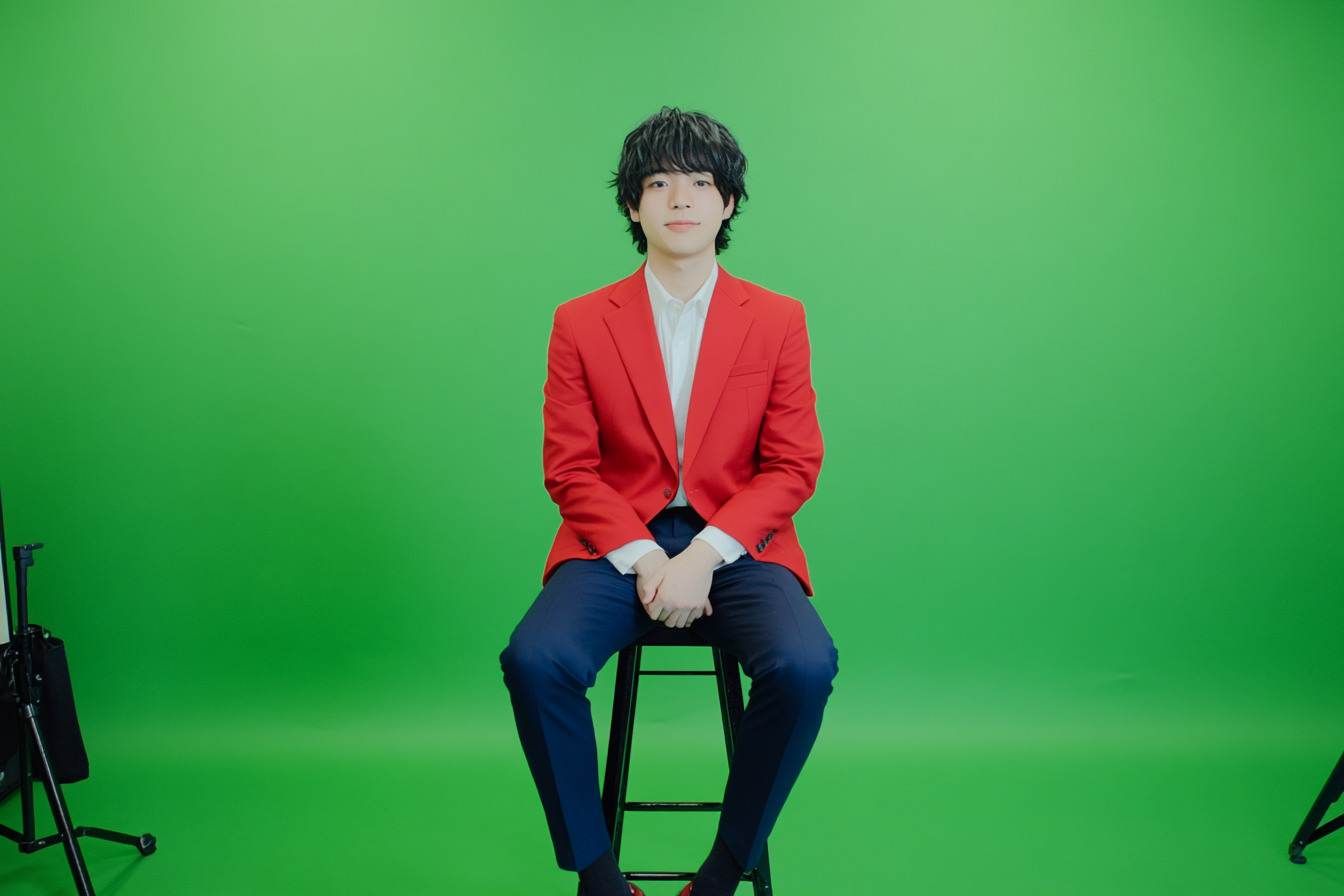 Japanese man in red blazer sitting on stool.