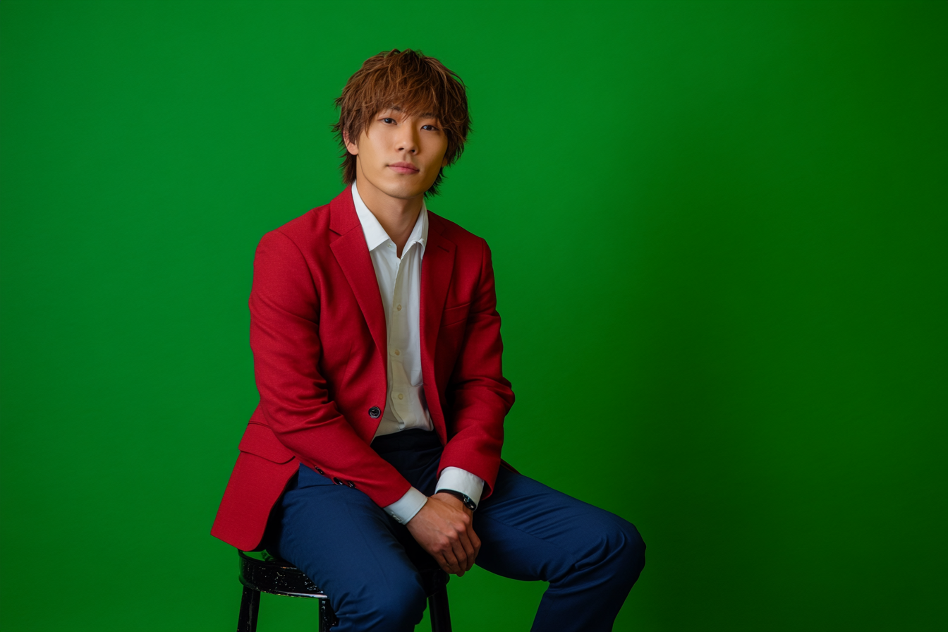 Japanese man in red blazer sitting on stool.