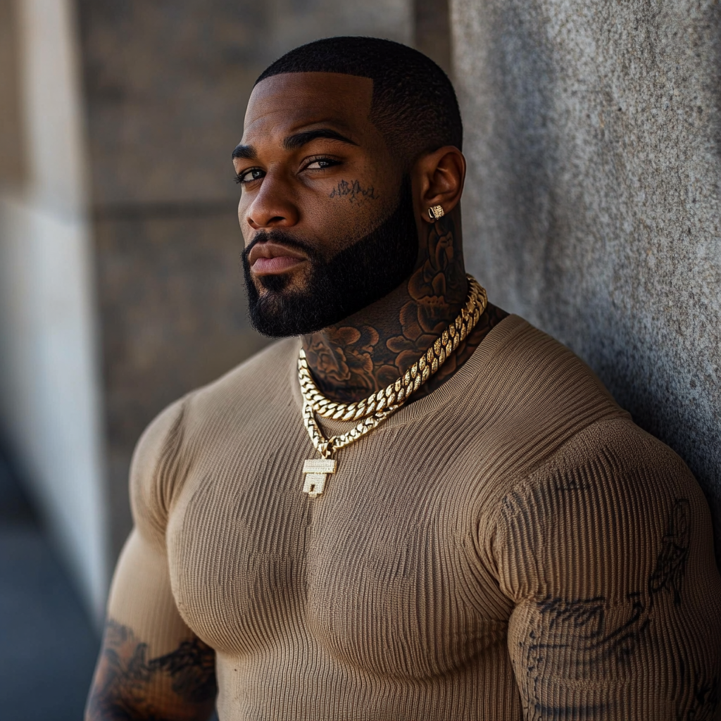 Tattooed black man with dark skin, Cuban chain & beard among a wall.