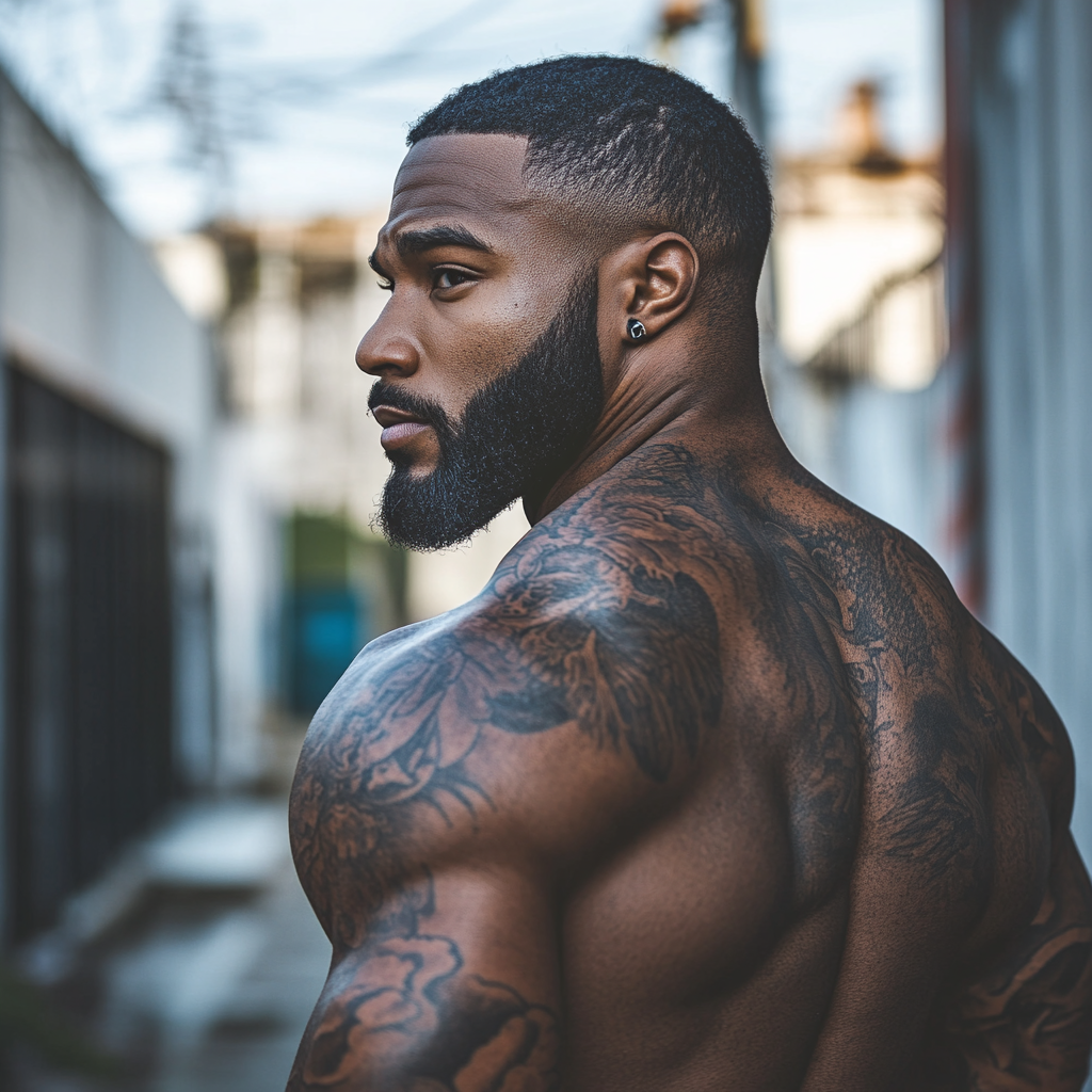 Dark skinned man with tattoos stands outside.