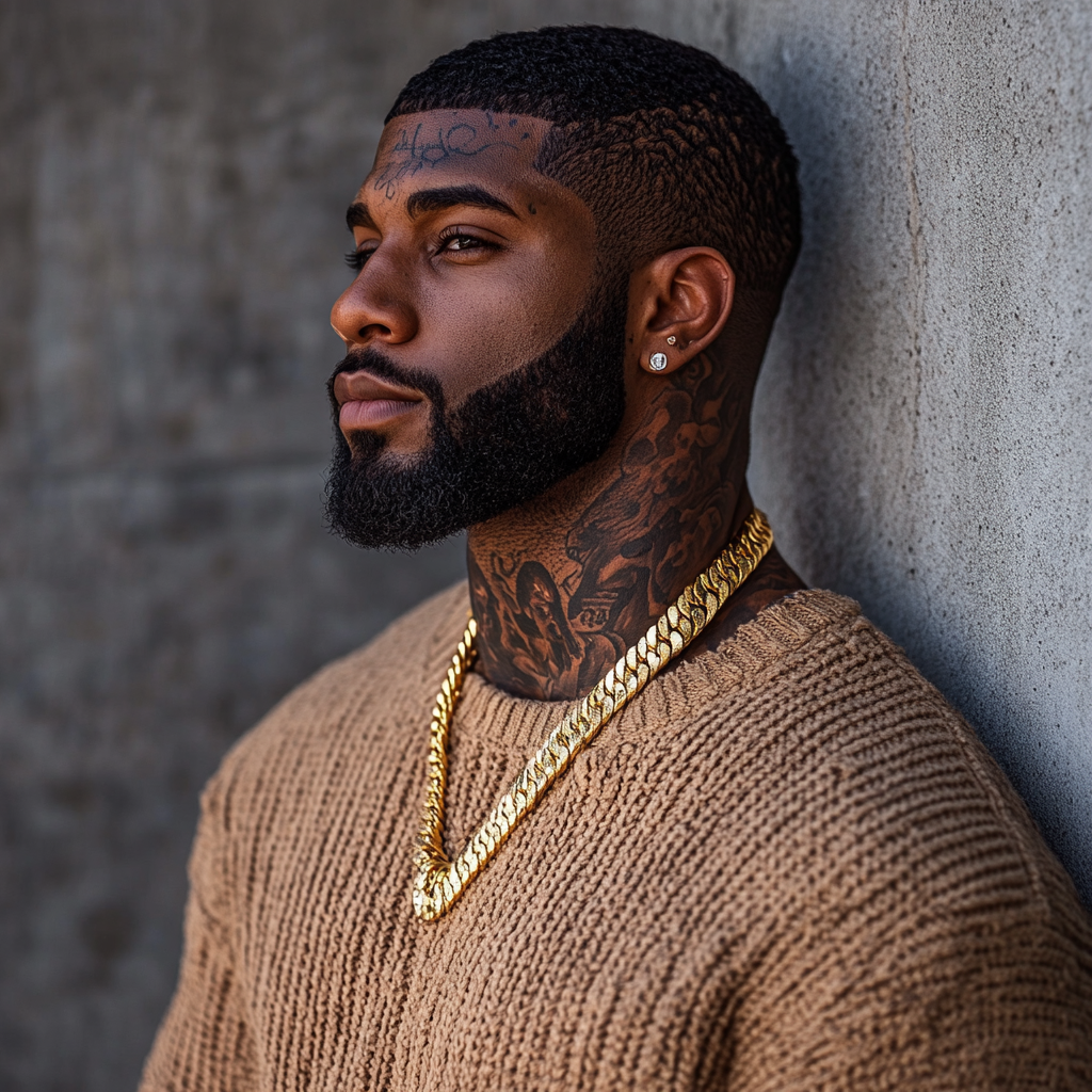 Tattooed black man with dark skin, Cuban chain & beard among a wall.